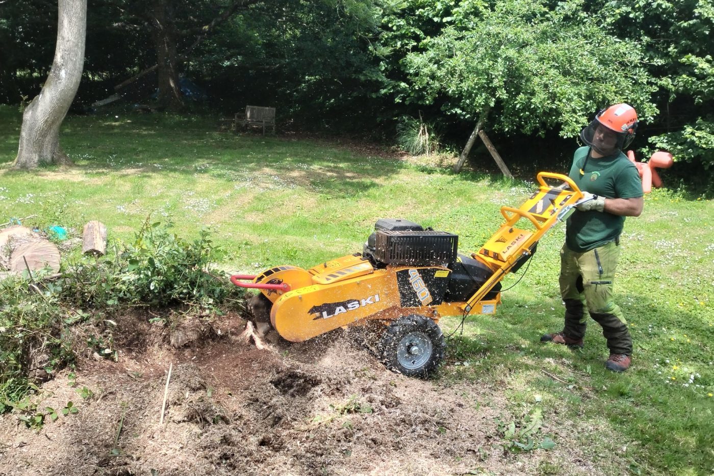Person operating a yellow stump grinder machine to remove a tree stump in a grassy garden area, wearing protective gear including a helmet and ear protection.